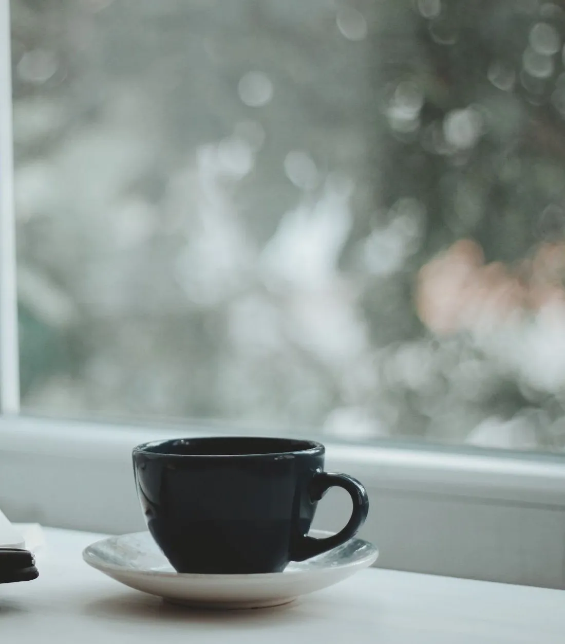 A dark cup on a saucer by a rain-speckled window