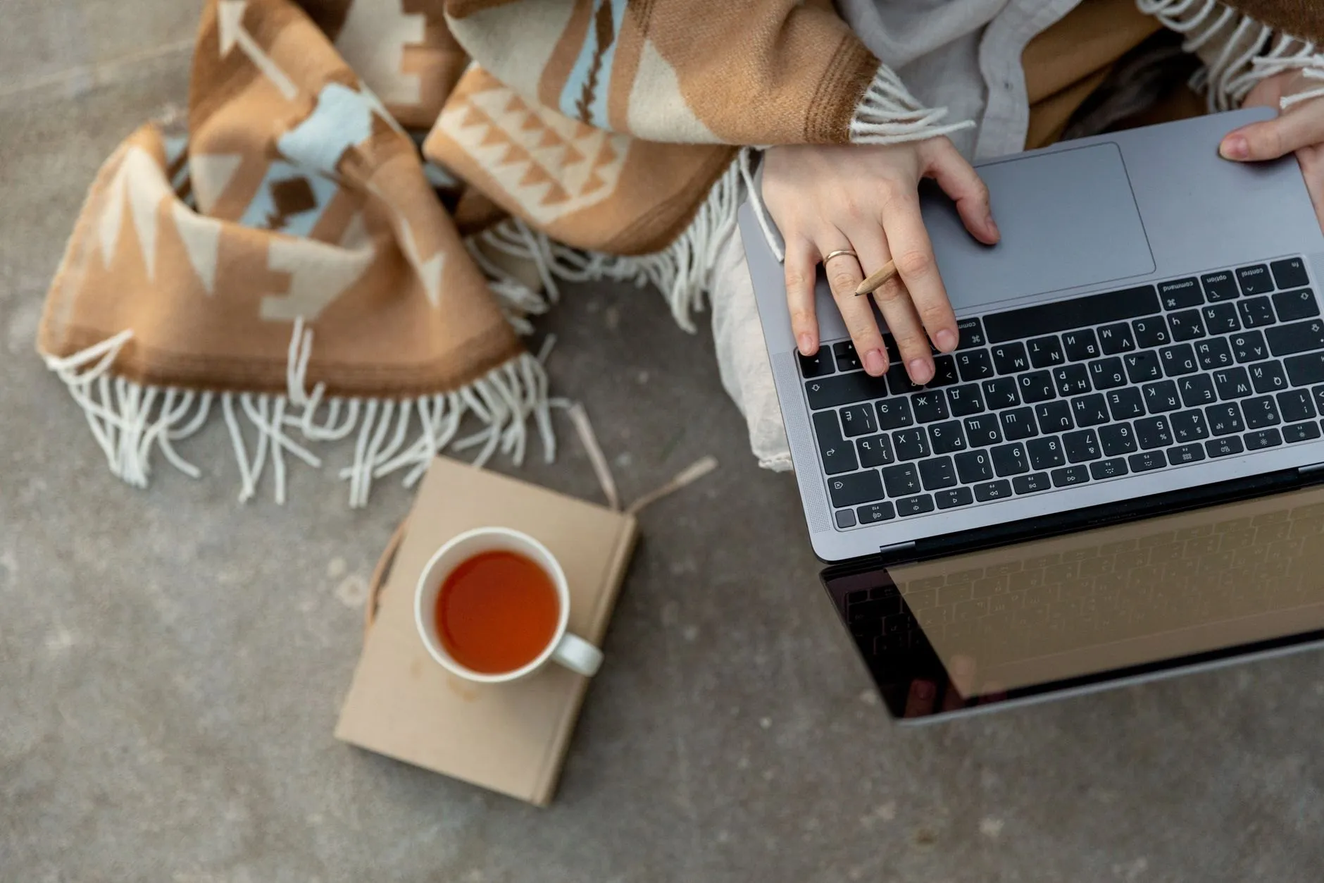 Hands on a laptop keyboard beside a cup of tea and a notebook, seen from above