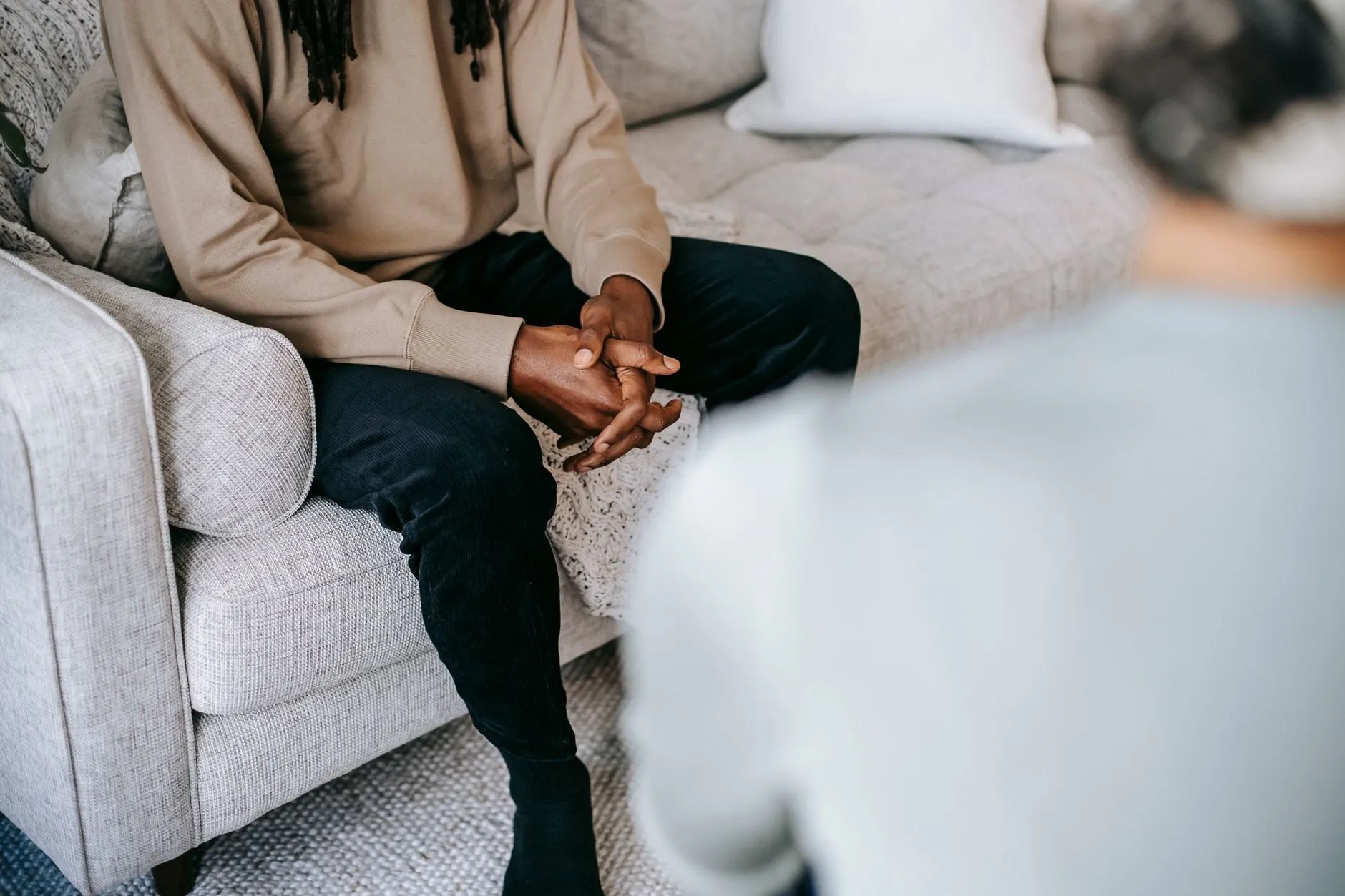 A client sitting on a pale sofa with hands clasped, a therapist out of focus in the foreground