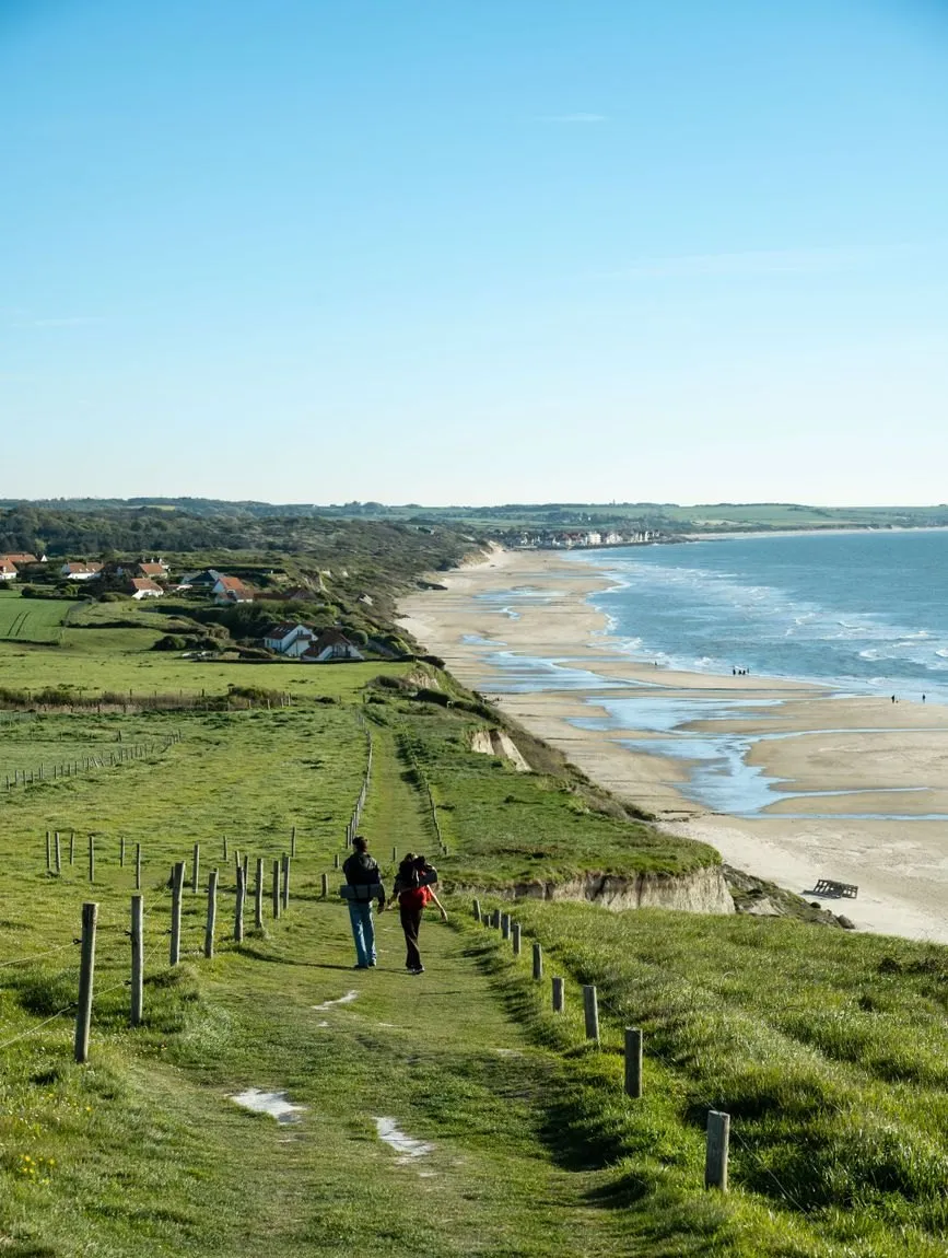 Two people walking away along a grassy clifftop path above a long sandy beach